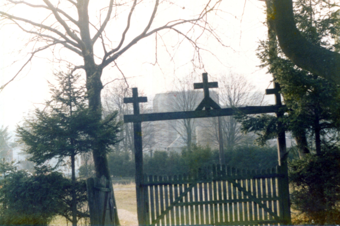 Röm. kath. Friedhof: Eingangsbereich mit Holztor, auf dessen oberem Querbalken drei Kreuzen aufgesetzt sind